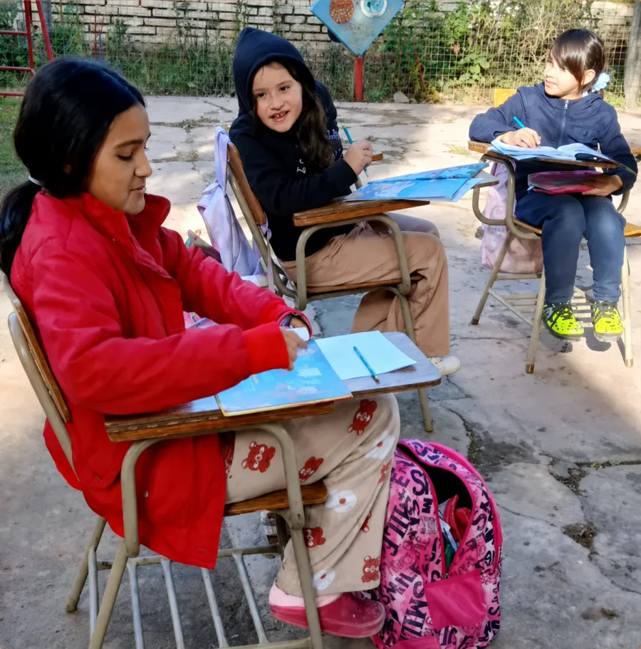 Tres chicas estudian en sus pupitres, una levanta la vista y sonríe.
