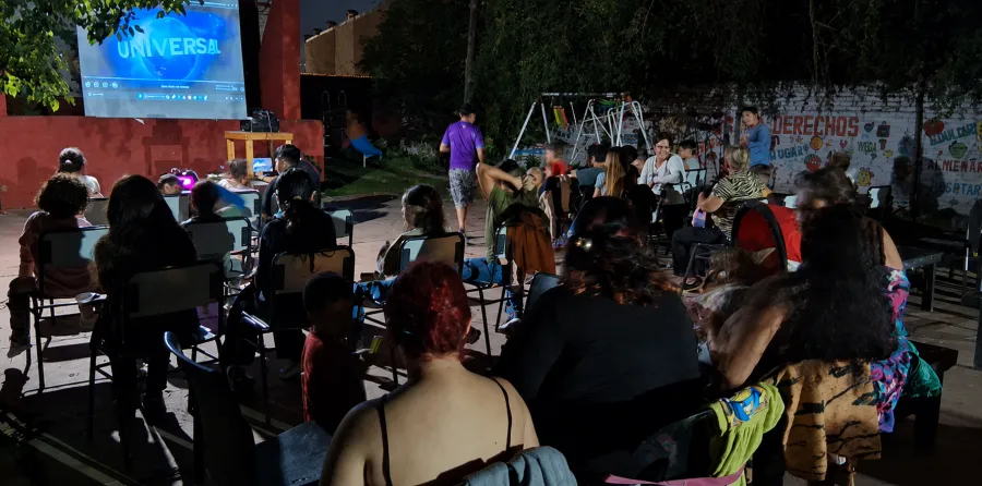 El barrio sentado en sillas en el patio de noche, mirando la película proyectada en la pared.