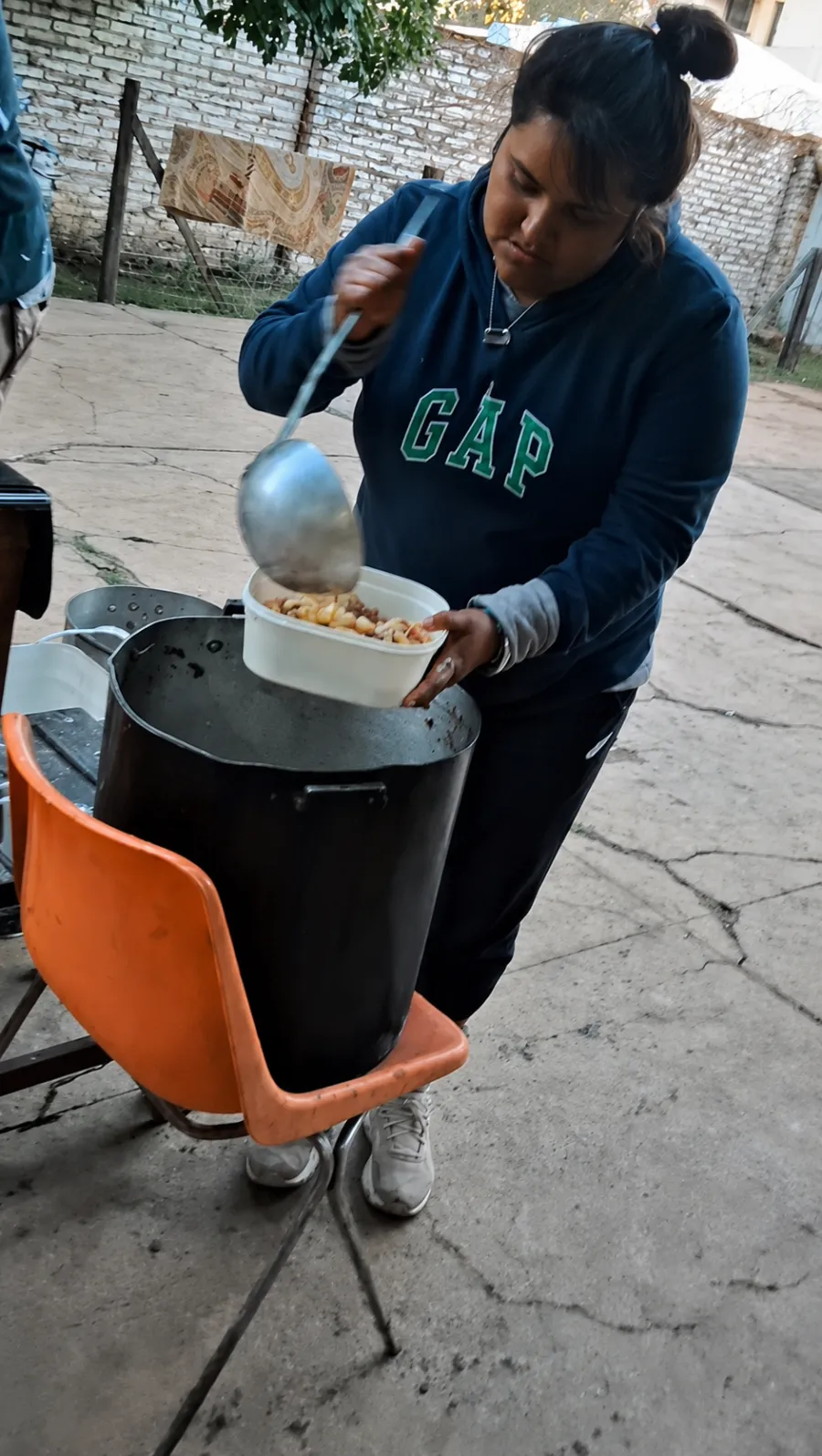 Una vecina sirve con cucharón una porción de guiso en un tupper, al lado de la olla del comedor.